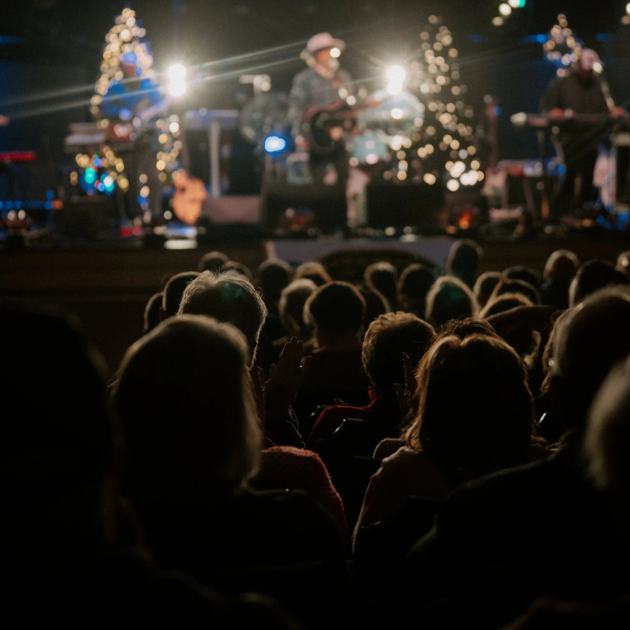 A group of musicians performing on stage at a concert, with Christmas trees and bright lights in the background.