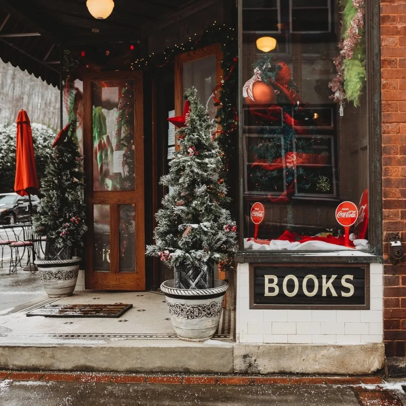 A storefront decorated with Christmas ornaments, with two snow-dusted Christmas trees in large planters outside. The window display includes festive wreaths and red Coca-Cola signs, with the word 'BOOKS' on a sign at the bottom.