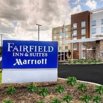 Signboard for Fairfield Inn & Suites Marriott hotel in front of a modern building with an American flag, surrounded by green plants and parking area.
