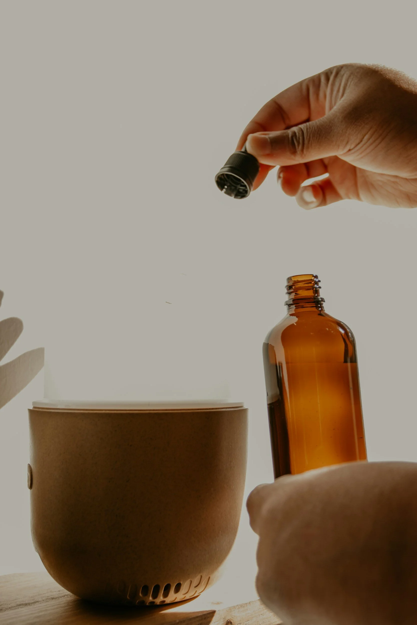 A person holding an essential oil bottle over a diffuser, with a brown container on a wooden surface.