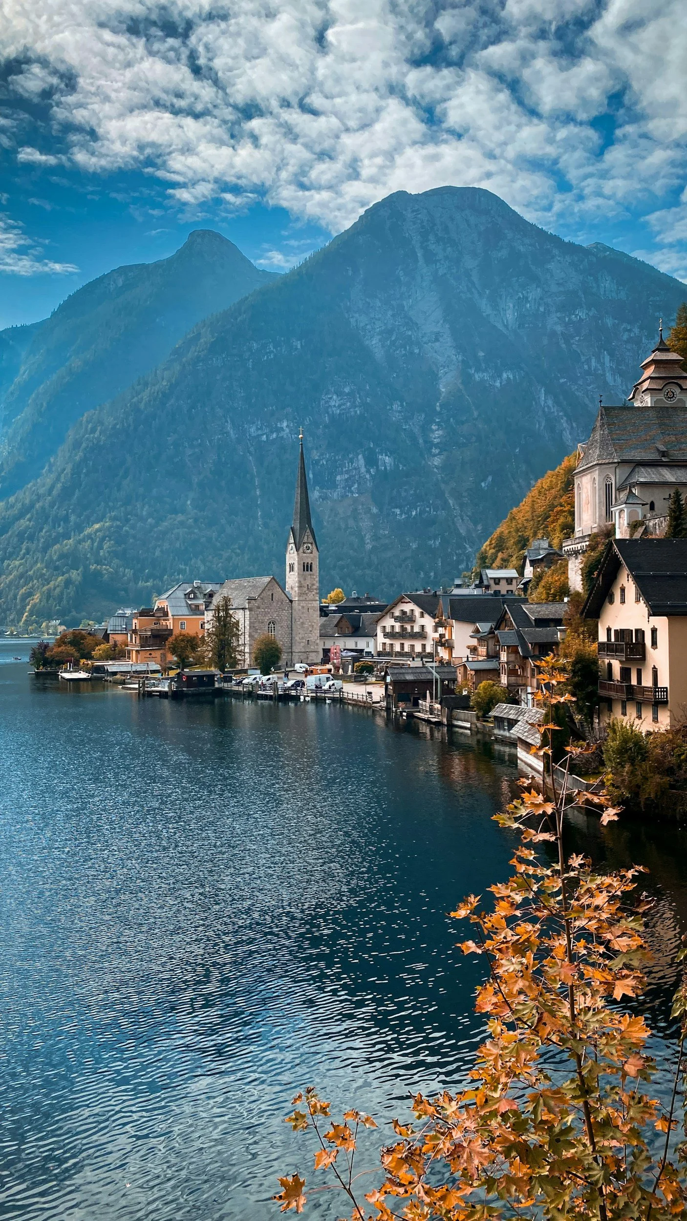 A picturesque lakeside village with colorful houses, a church with a tall steeple, and mountain peaks in the background, and autumn foliage in the foreground.