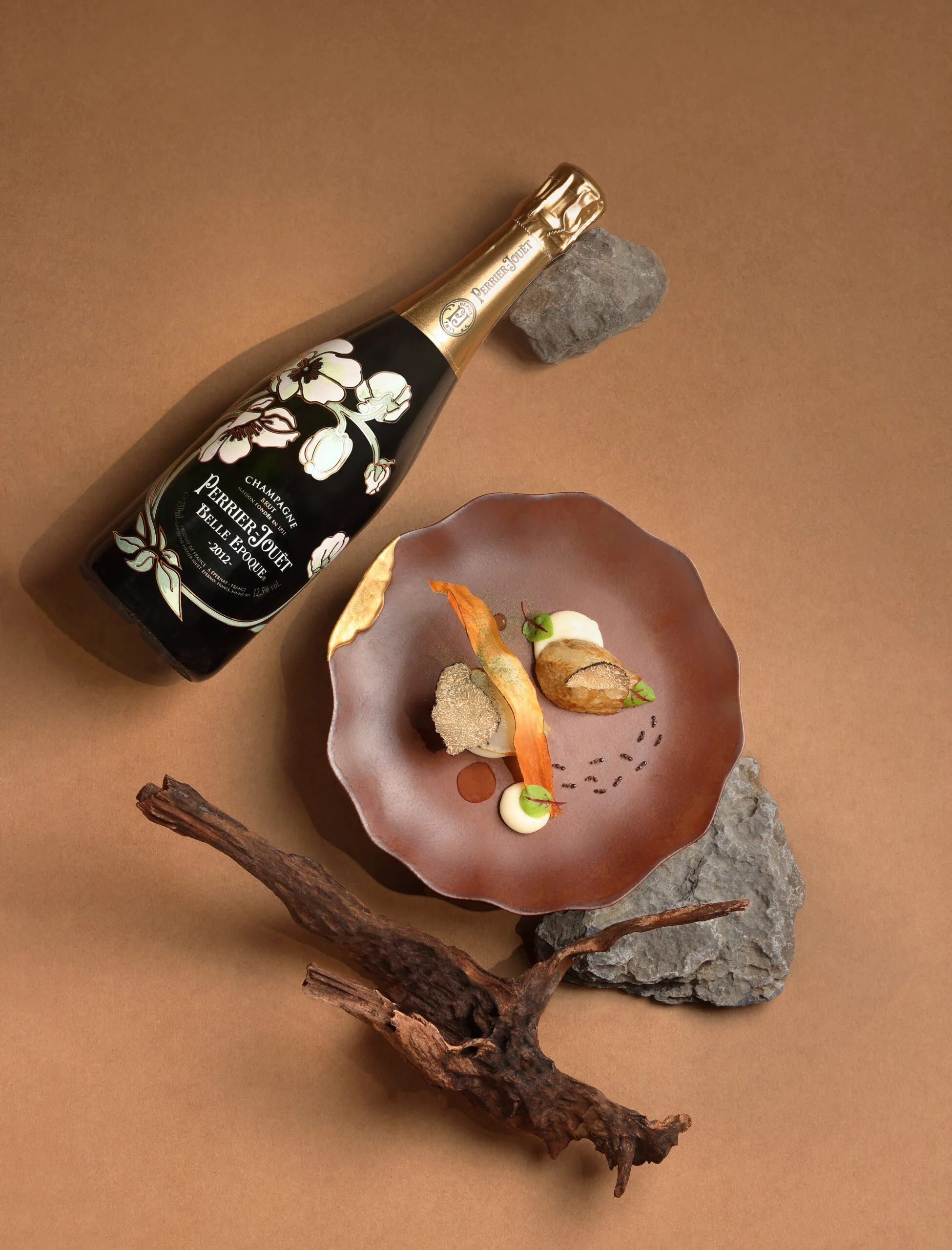 A bottle of Perrier-Jouët champagne, two rocks, a brown dish with three gourmet food items garnished with microgreens and sauces, and a piece of driftwood on a brown background.