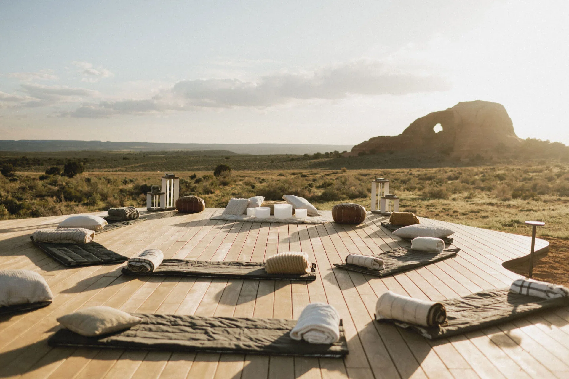 A meditation setup on a wooden platform in an open desert landscape with a large rock formation and blue sky in the background, including pillows, rolled towels, candles, and lanterns arranged in a circle.