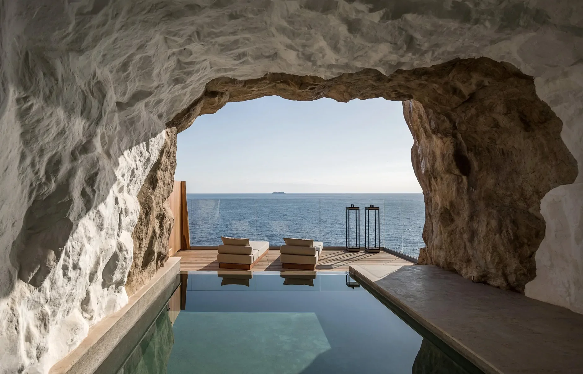 View of the ocean through a cave-like opening with two lounge chairs and lanterns on a wooden deck.