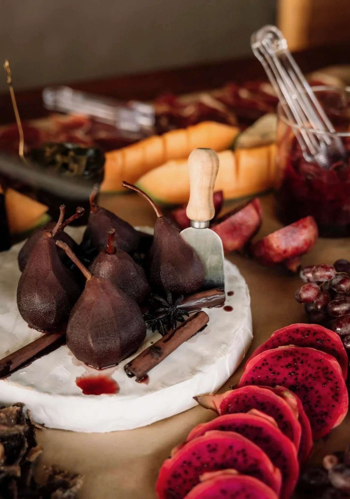 Assorted fresh fruits including sliced dragon fruit, red grapes, pear, peach, and figs displayed around a cheese platter with a cheese knife.