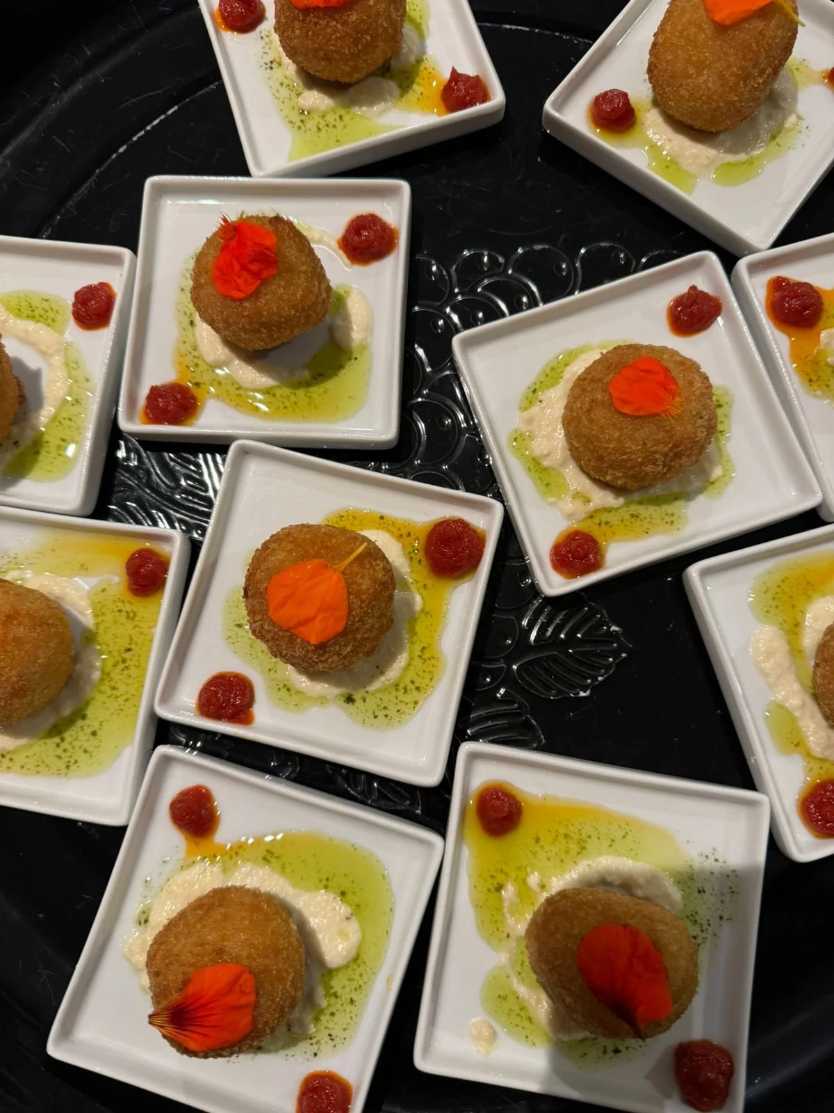 Several small white square plates each holding a breaded fried appetizer garnished with a red edible flower, with green and white sauces and small dollops of red sauce around them.