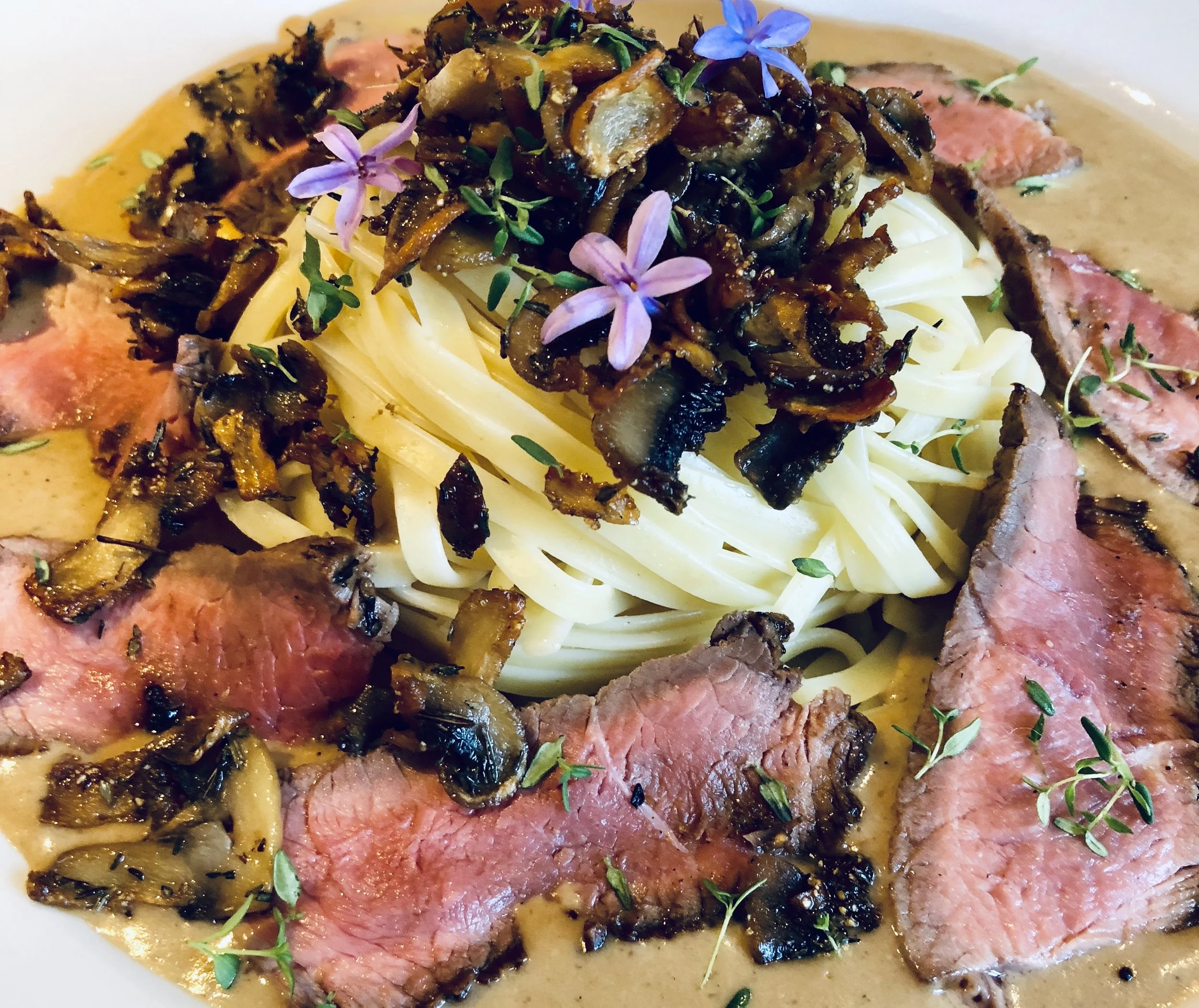 A plate with pasta topped with caramelized onions, slices of medium-rare steak, and garnished with herbs and edible flowers.