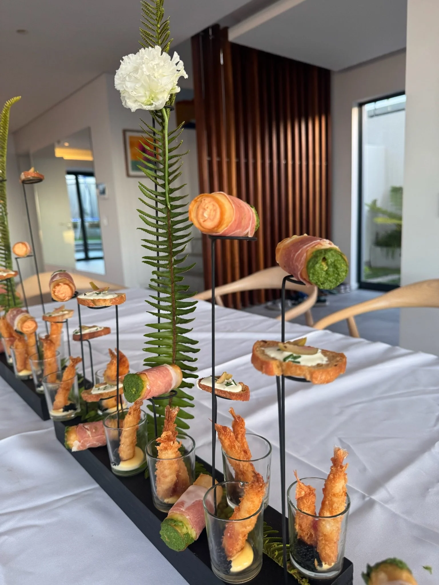 Elevated appetizer display with skewers of bacon-wrapped green vegetables, slices of bread topped with cream and herbs, and breaded fried shrimp, arranged on a black tray with green fern accents.