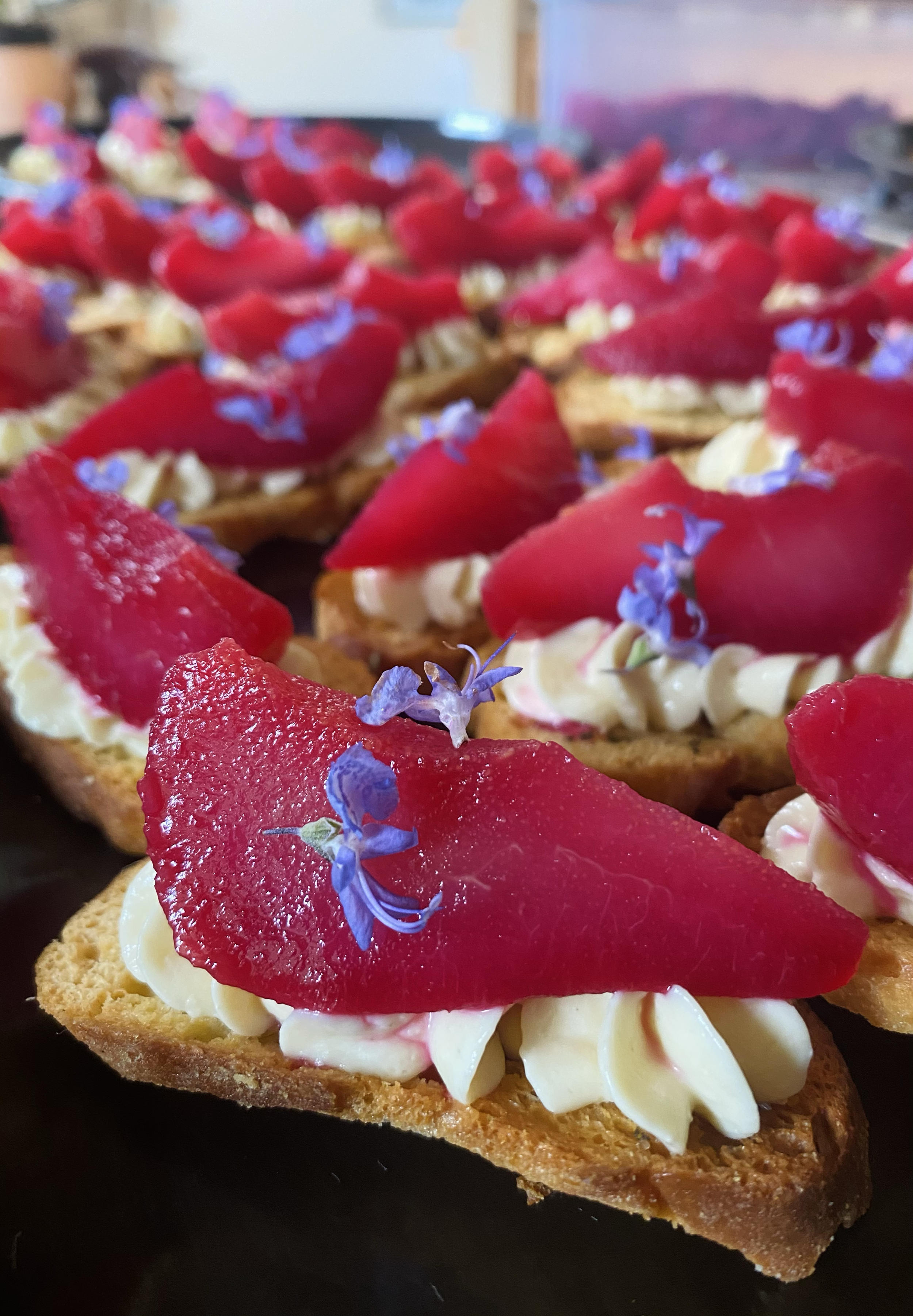 Close-up of toasted bread slices topped with cream, red fruit slices, and small purple edible flowers.