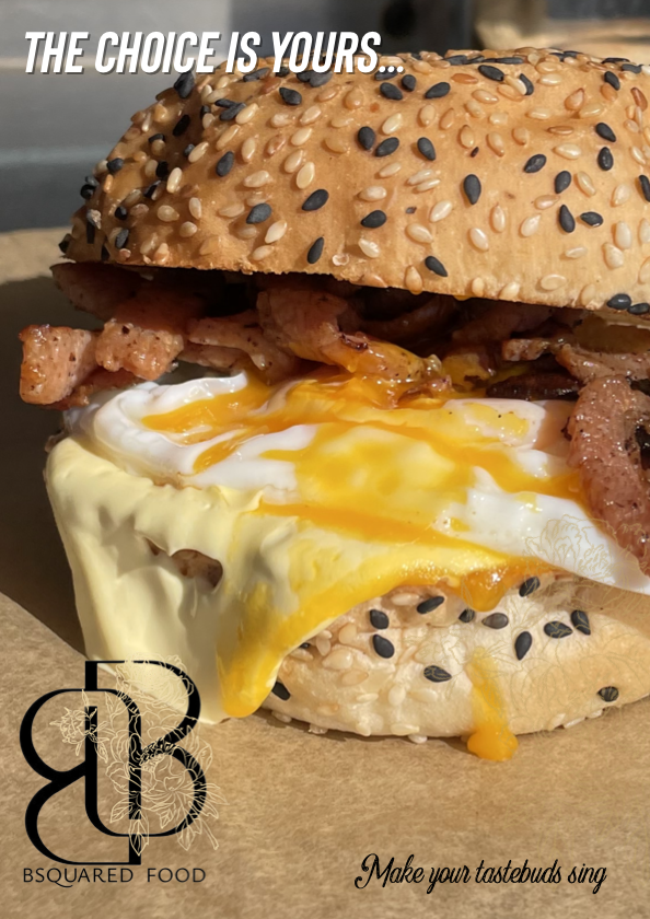 A sesame seed sandwich filled with crispy bacon and a fried egg with runny yolk, topped with text that says 'The choice is yours...' and a logo for BSQUARED FOOD with the tagline 'Make your taste buds sing'.