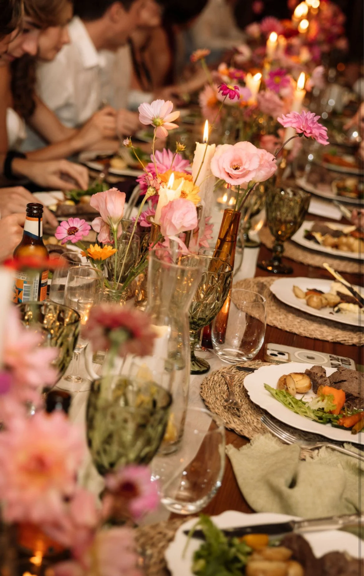 A festive dinner table with floral centerpieces, lit candles, and plates of food, surrounded by people.