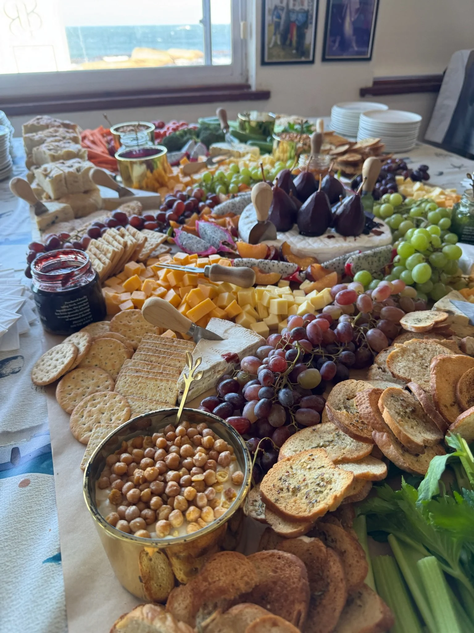 A large breakfast or brunch spread featuring various cheeses, crackers, grapes, figs, sliced bread, chickpeas in a bowl, and jars of jam, with a window and framed pictures in the background.