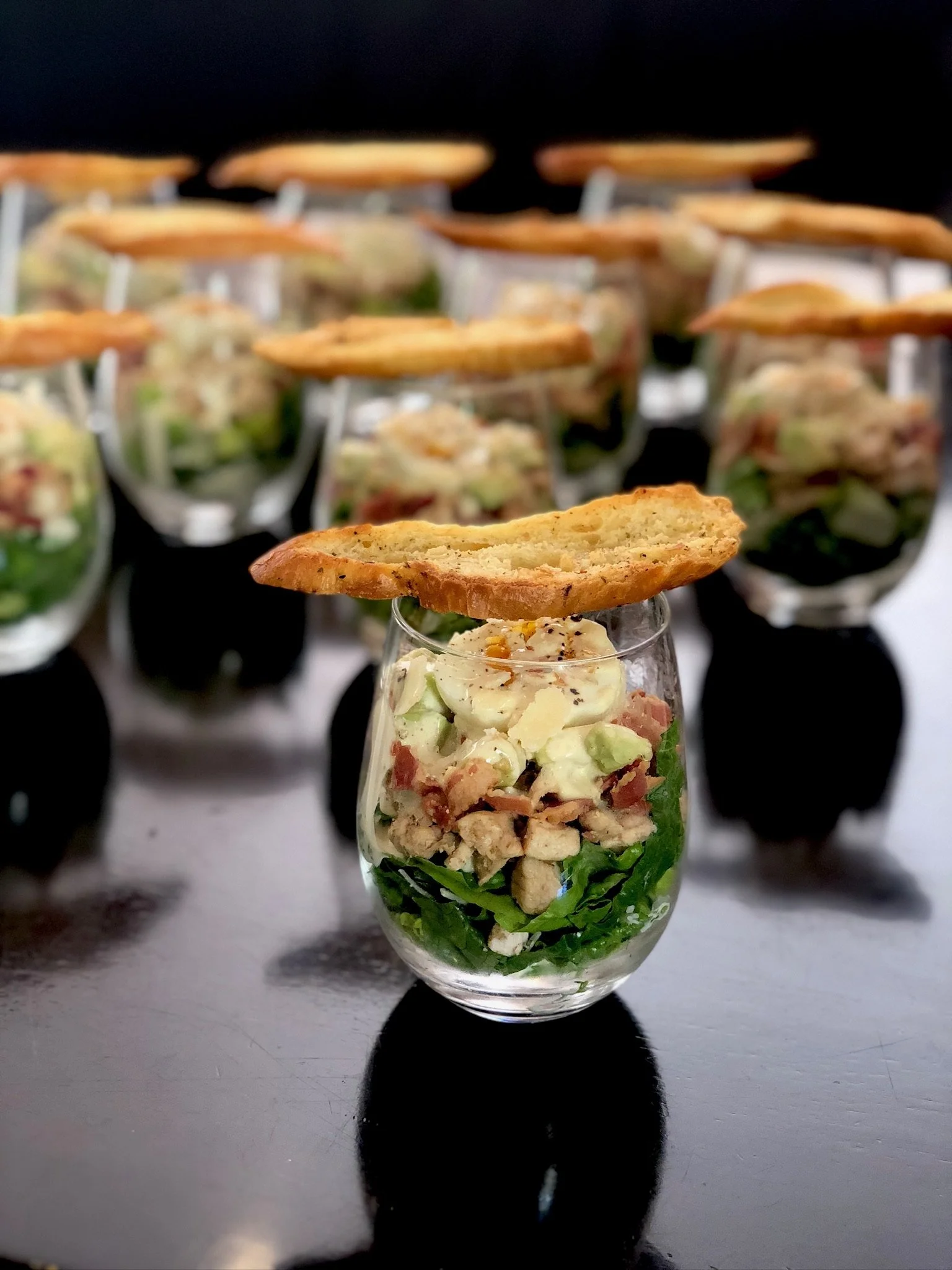 Individual servings of layered chicken caesar salad with a slice of toasted bread on top, arranged on a dark surface.