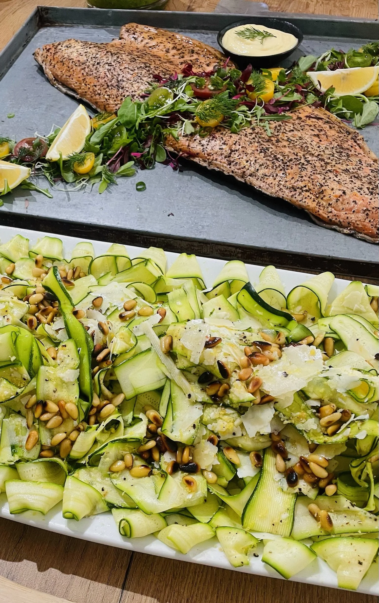 Grilled salmon fillets with side salad, lemon wedges, and dipping sauce on a metal tray, with zucchini and pine nuts salad in the foreground.