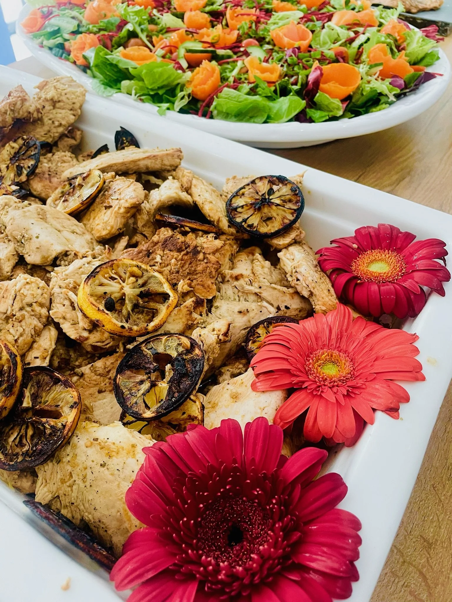 A serving platter of grilled chicken topped with grilled lemon slices, decorated with bright pink and red flowers. In the background, there is a large salad bowl filled with mixed greens, shredded carrots, and other vegetables.
