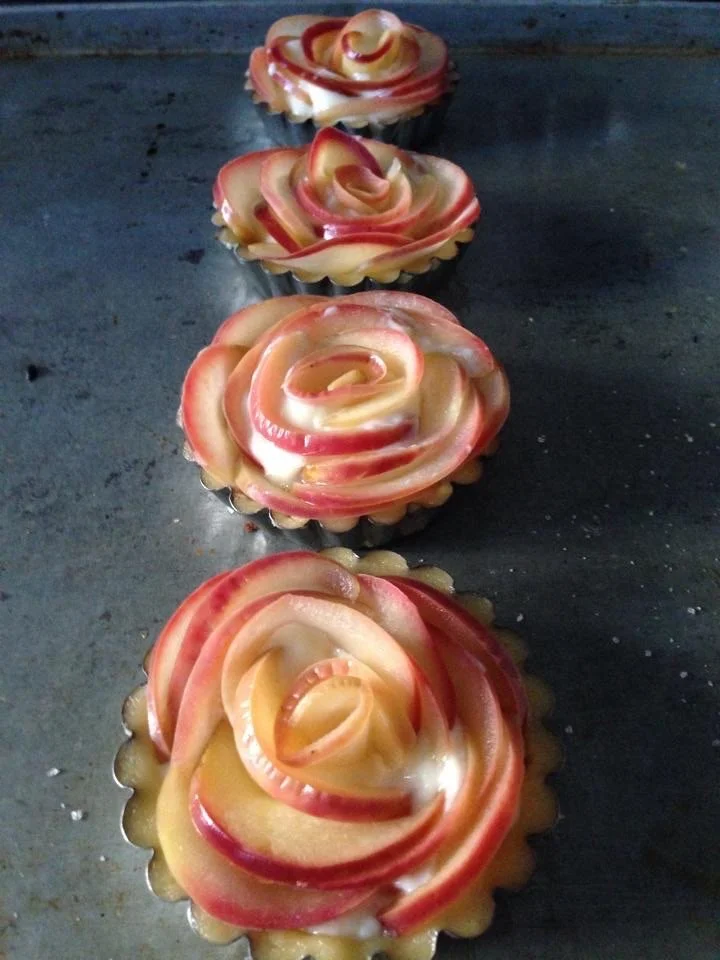 Four apple rose tarts arranged in a row on a dark baking sheet, each tart topped with thin apple slices arranged in a rose pattern.
