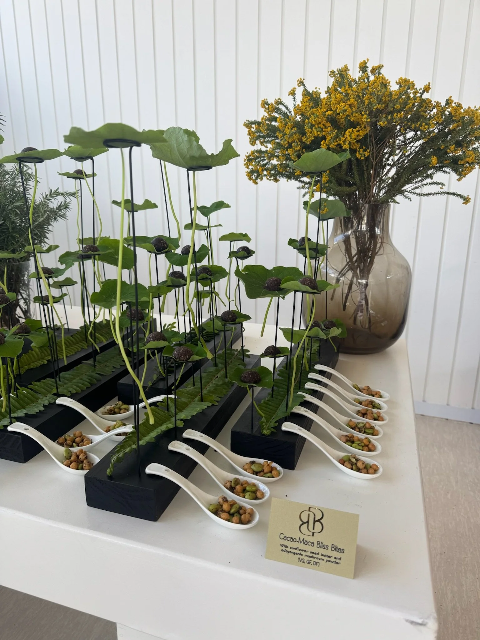Display of small bites called Cacao-Maca Bliss Bites on a white table with small white spoons containing the bites, set against a backdrop of green plants, a large vase with yellow flowers, and a small sign describing the bites with ingredients inclu