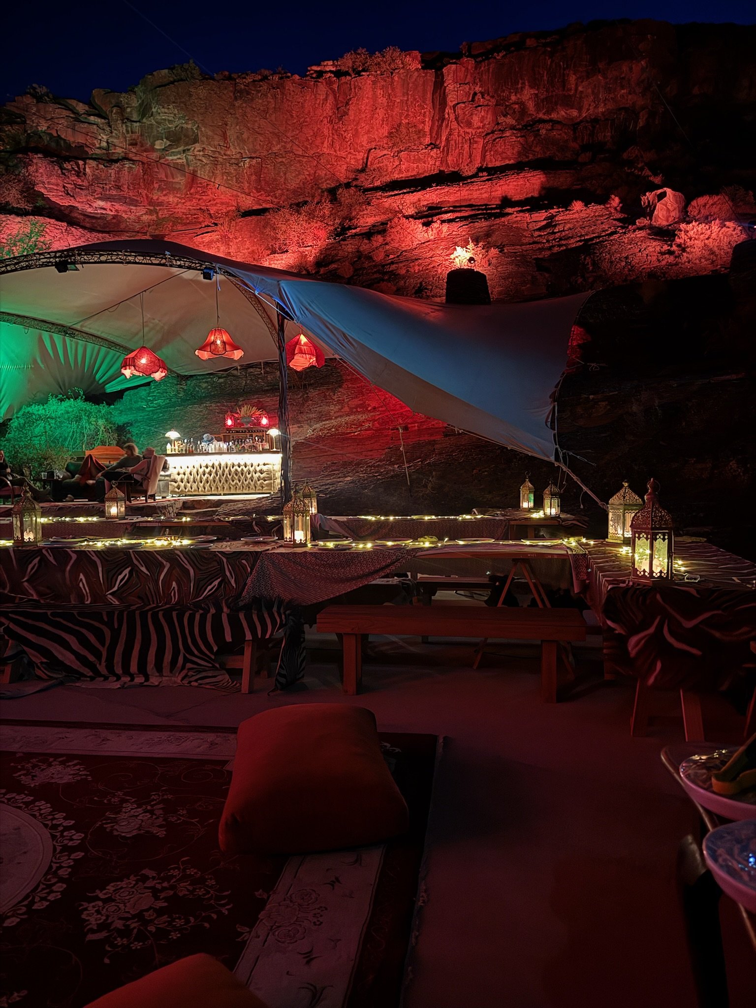 A nighttime outdoor lounge area with a rock cliff illuminated in red in the background, covered by a canopy with hanging red lamps, decorated with lanterns and string lights, containing seating and a bar area.