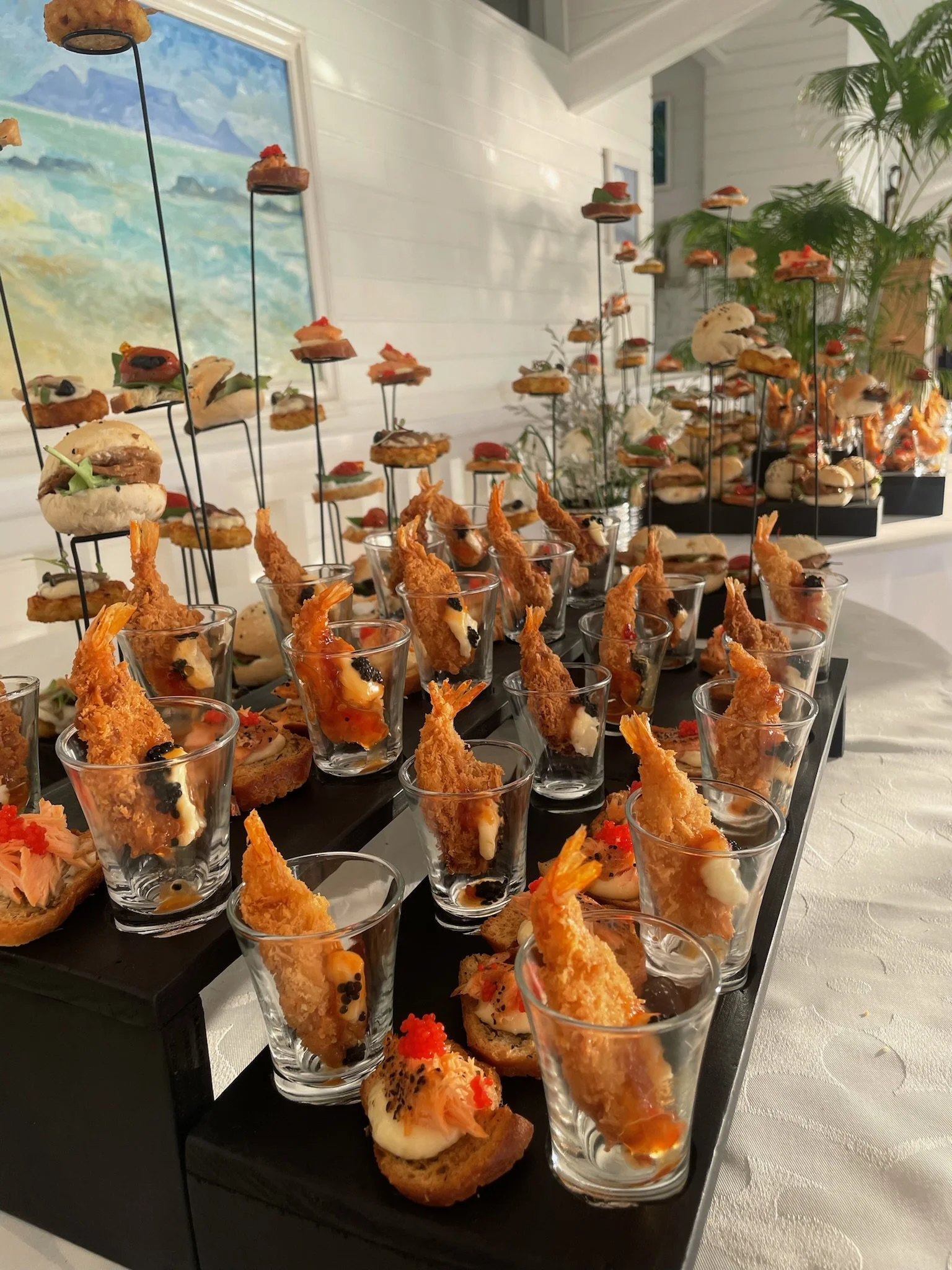 Assorted gourmet appetizers on display, including mini sandwiches and fried shrimp, arranged on black tiered trays with a white tablecloth and decorative plants in the background.