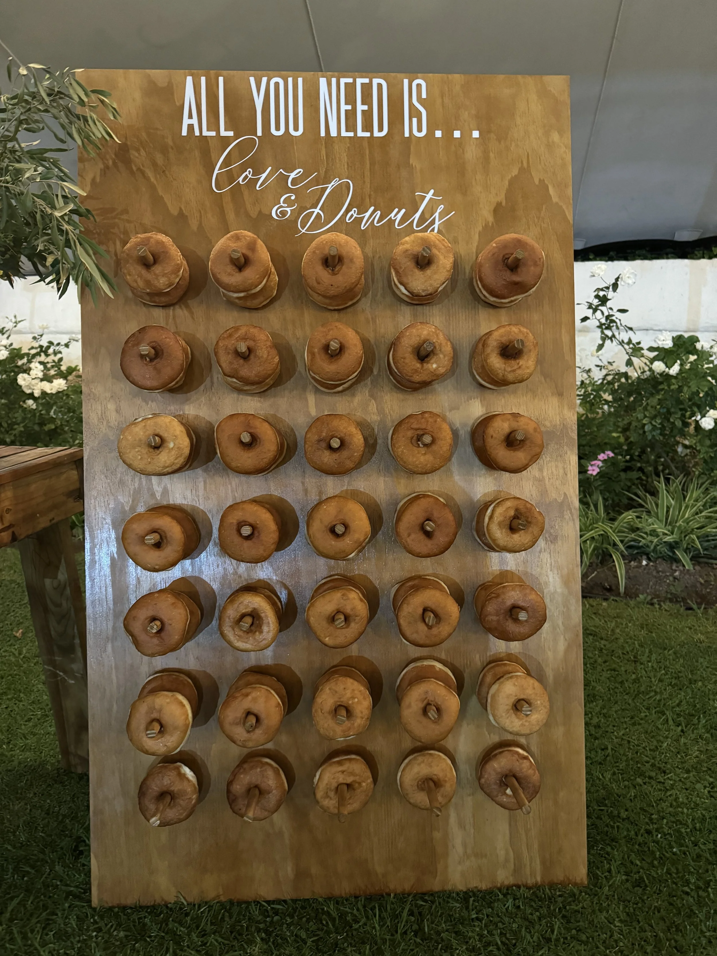 Wooden display board with multiple donuts on sticks, arranged in rows. A sign at the top reads, 'All you need is... love & donuts'.