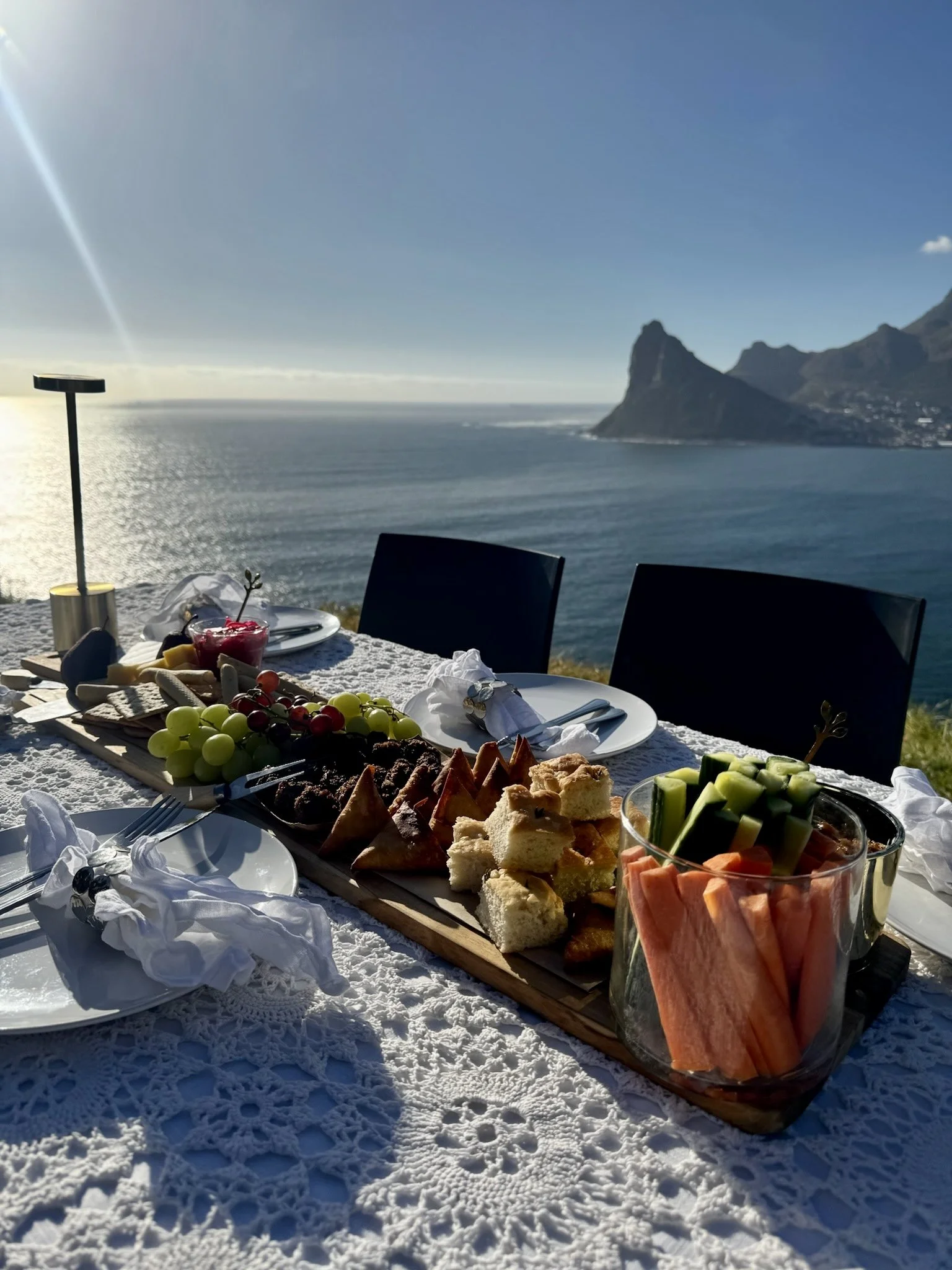 An outdoor table set with a white lace tablecloth, holding a cheese platter, fresh grapes, and a bowl of sliced cucumbers and carrots, overlooking a coastal landscape with mountains and the ocean during sunset.