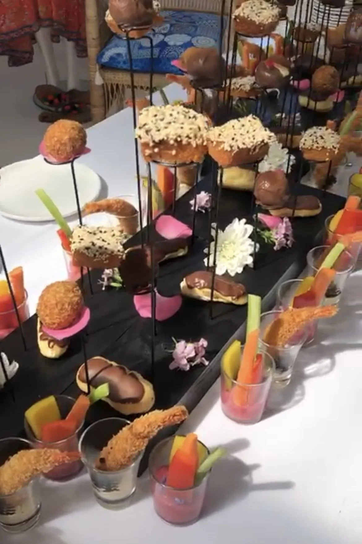 A display of assorted savory and sweet appetizers including croquettes, sandwiches, and dips garnished with vegetables, arranged on a black standing tray and small cups with vegetable sticks, decorated with flowers.