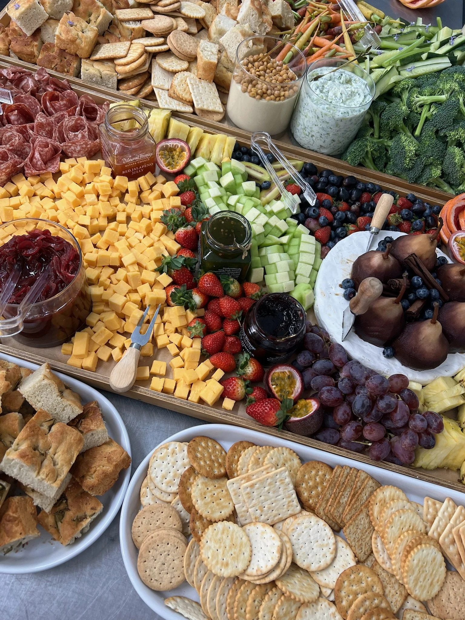 Assorted cheese, crackers, fresh vegetables, fruits, and dips on a platter, with bowls of bread and assorted snacks nearby.