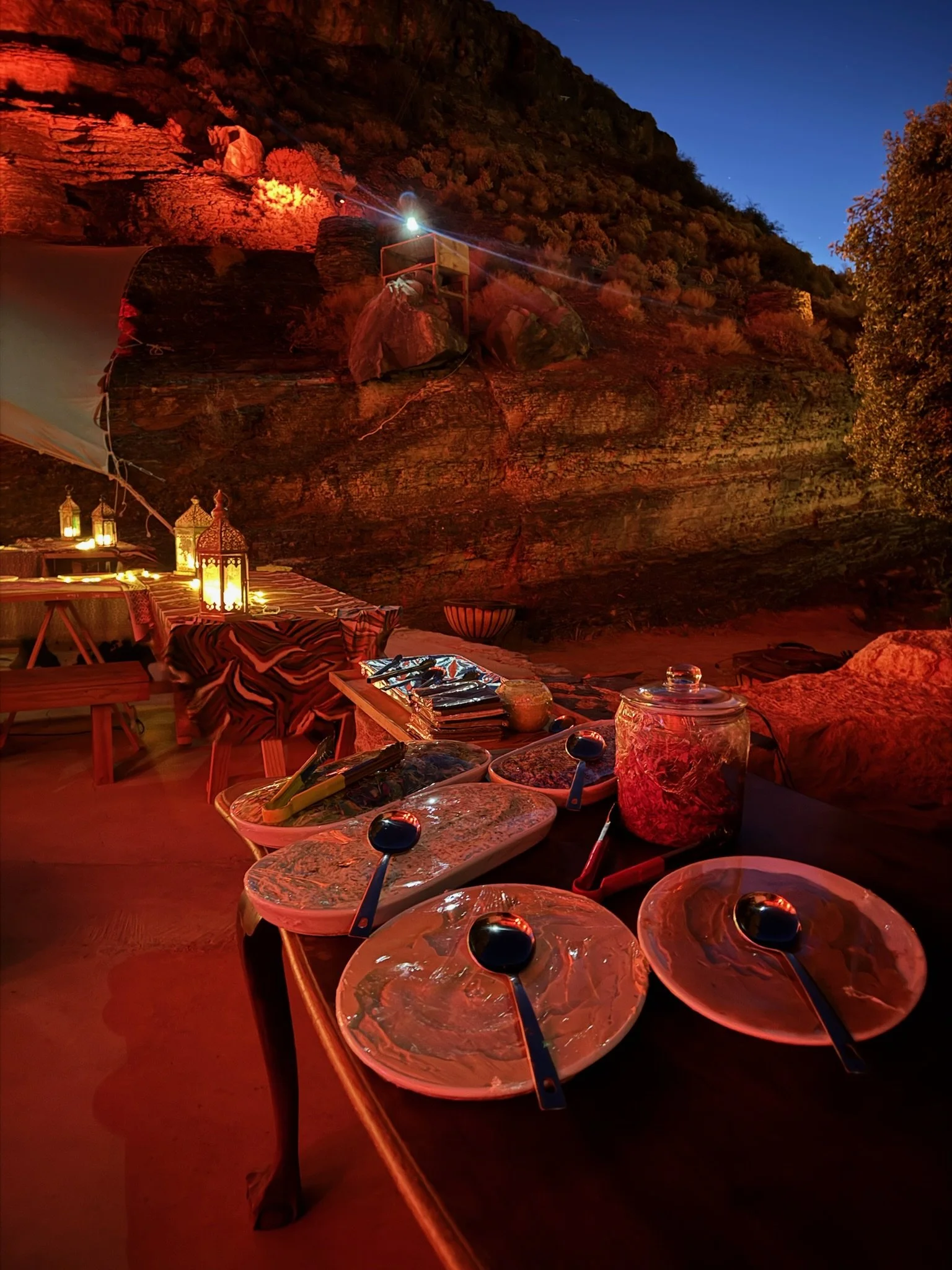 Outdoor dining setup with five empty plates with spoons, a glass jar, and various food items on a table covered with a patterned cloth. There is a rocky cliff in the background with a light shining on it, and lanterns and candles providing warm light