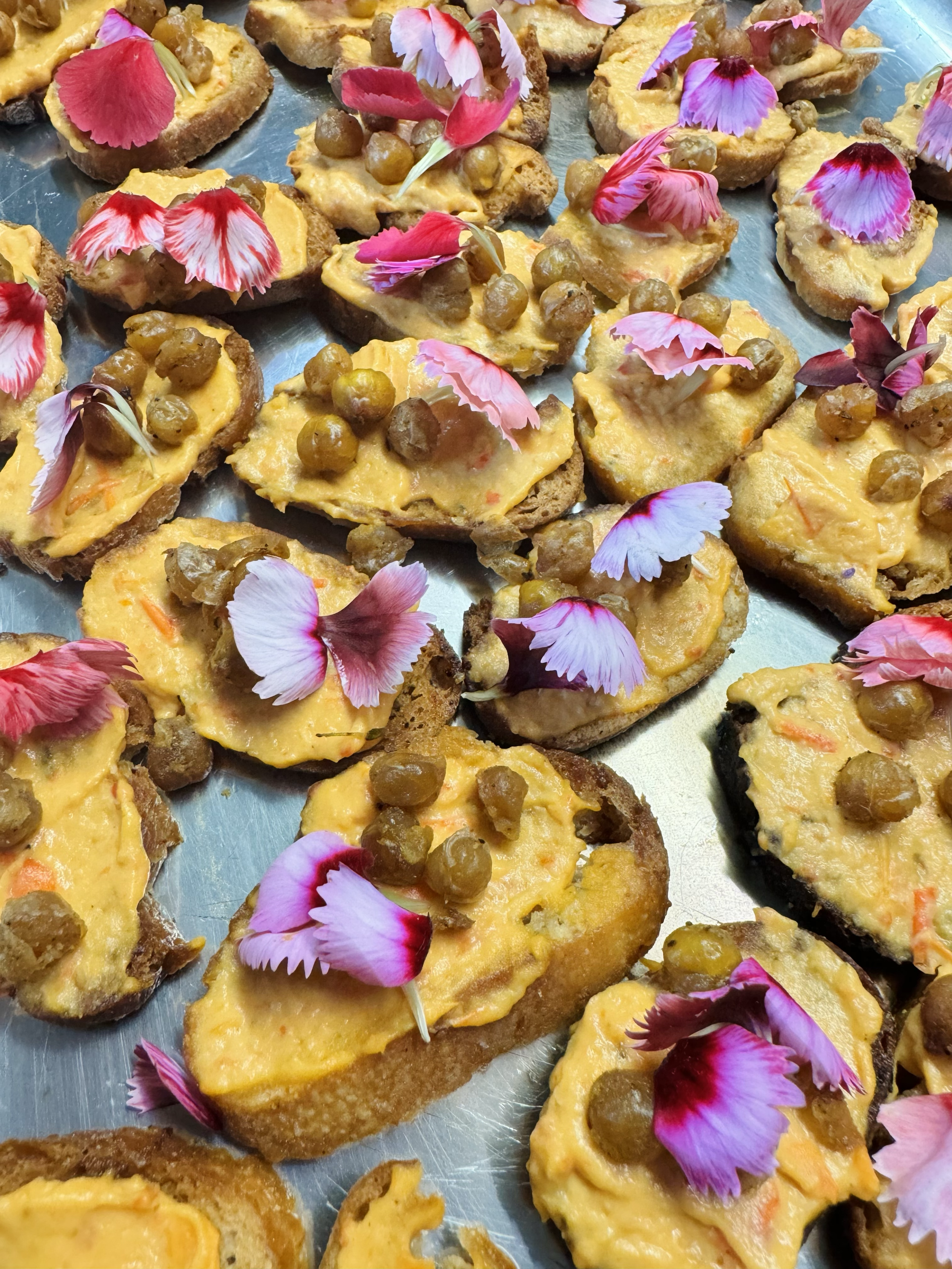 Close-up of small appetizers on a tray, featuring toasted bread slices topped with roasted red pepper humus  and roasted chickpeas garnished with pink and purple edible flower petals.