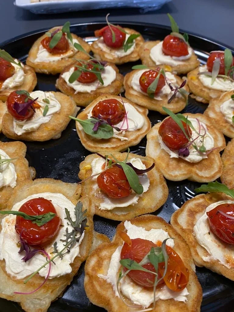 Mini toasted bread slices topped with cream cheese, cherry tomatoes, and microgreens, arranged on a black tray.