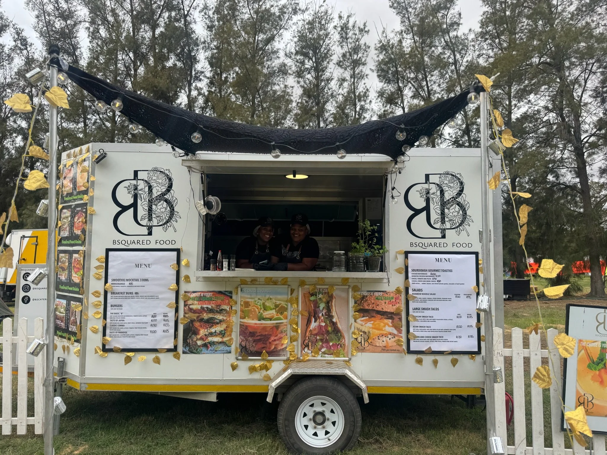 Food truck with a black awning, decorated with gold leaves and string lights, selling gourmet toast, salads, and smash tacos, with two smiling employees inside.