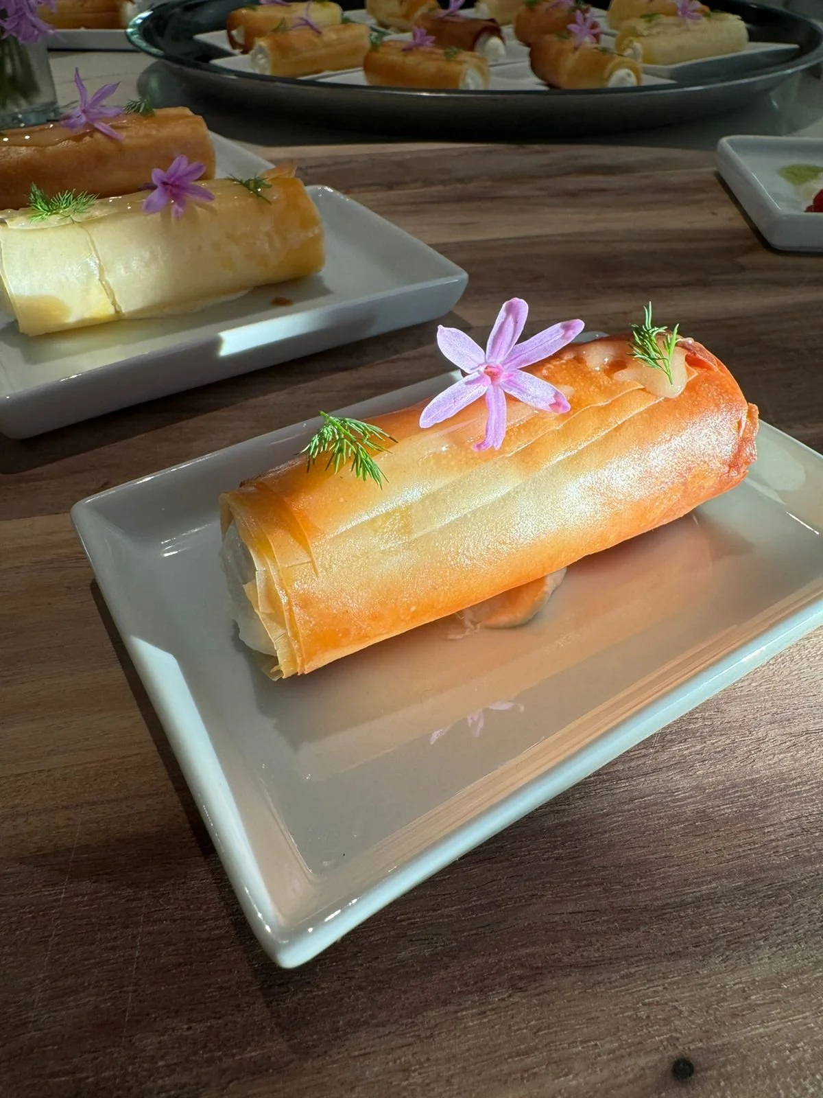 A plated, rolled, thin pastry with a pale orange color, garnished with purple flowers and small green herbs, served on a white rectangular plate with a wooden table surface. Additional similar pastries are visible on other plates in the background.
