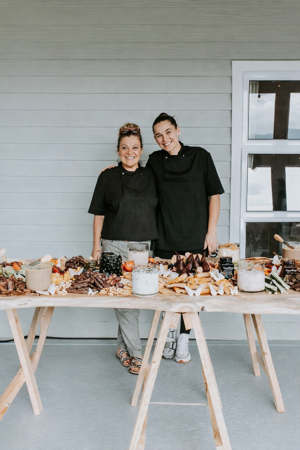 Two women in black chef uniforms standing behind a wooden table filled with various desserts, smiling indoors near a window.