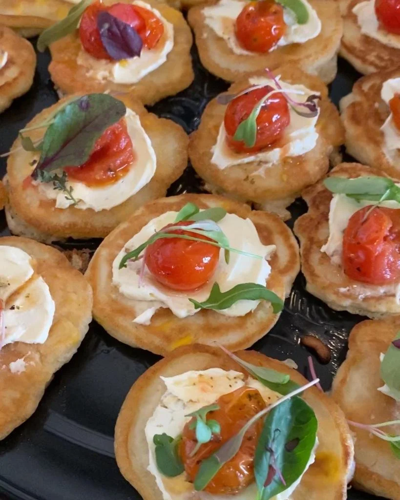 Close-up of bite-sized appetizers on a black platter, each consisting of a small toasted bread round topped with a dollop of cream or cheese, roasted cherry tomato, and garnished with microgreens or edible herbs.