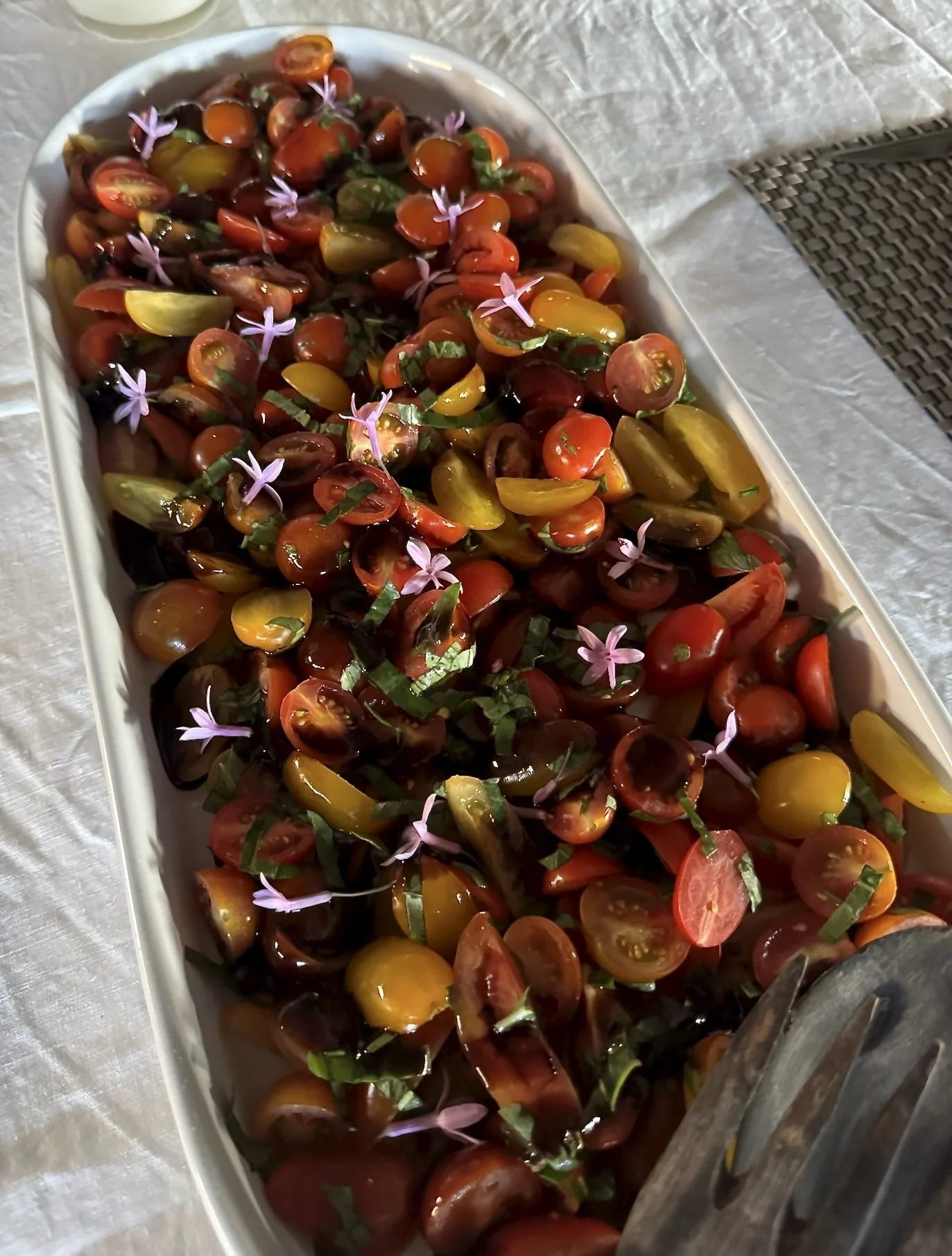 A large white serving dish filled with cherry tomato salad garnished with small purple flowers and chopped green herbs, placed on a white tablecloth.