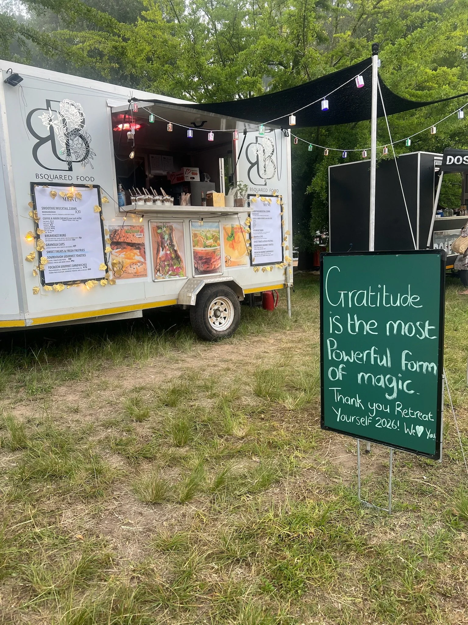 Food truck named BSquared Food with a menu, outdoor seating area with string lights, and a chalkboard sign with the quote 'Gratitude is the most powerful form of magic'.