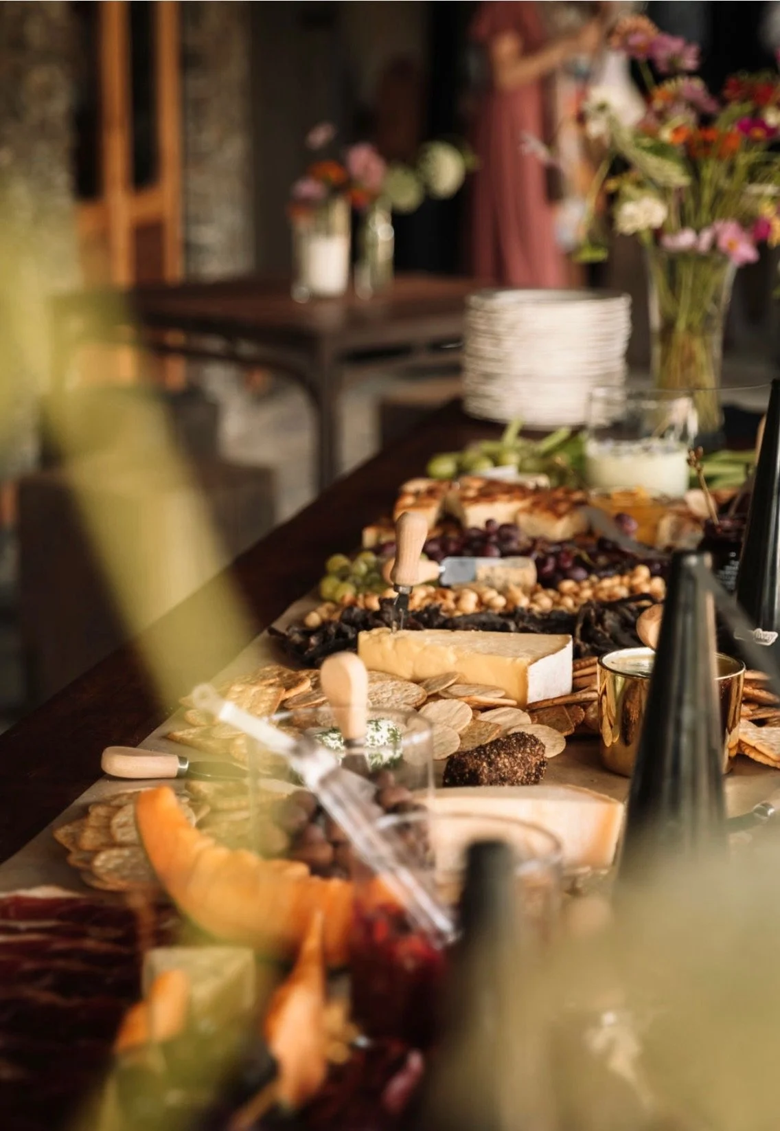A cheese and charcuterie spread on a wooden table with various cheeses, grapes, crackers, and dried fruits, with floral arrangements in the background.