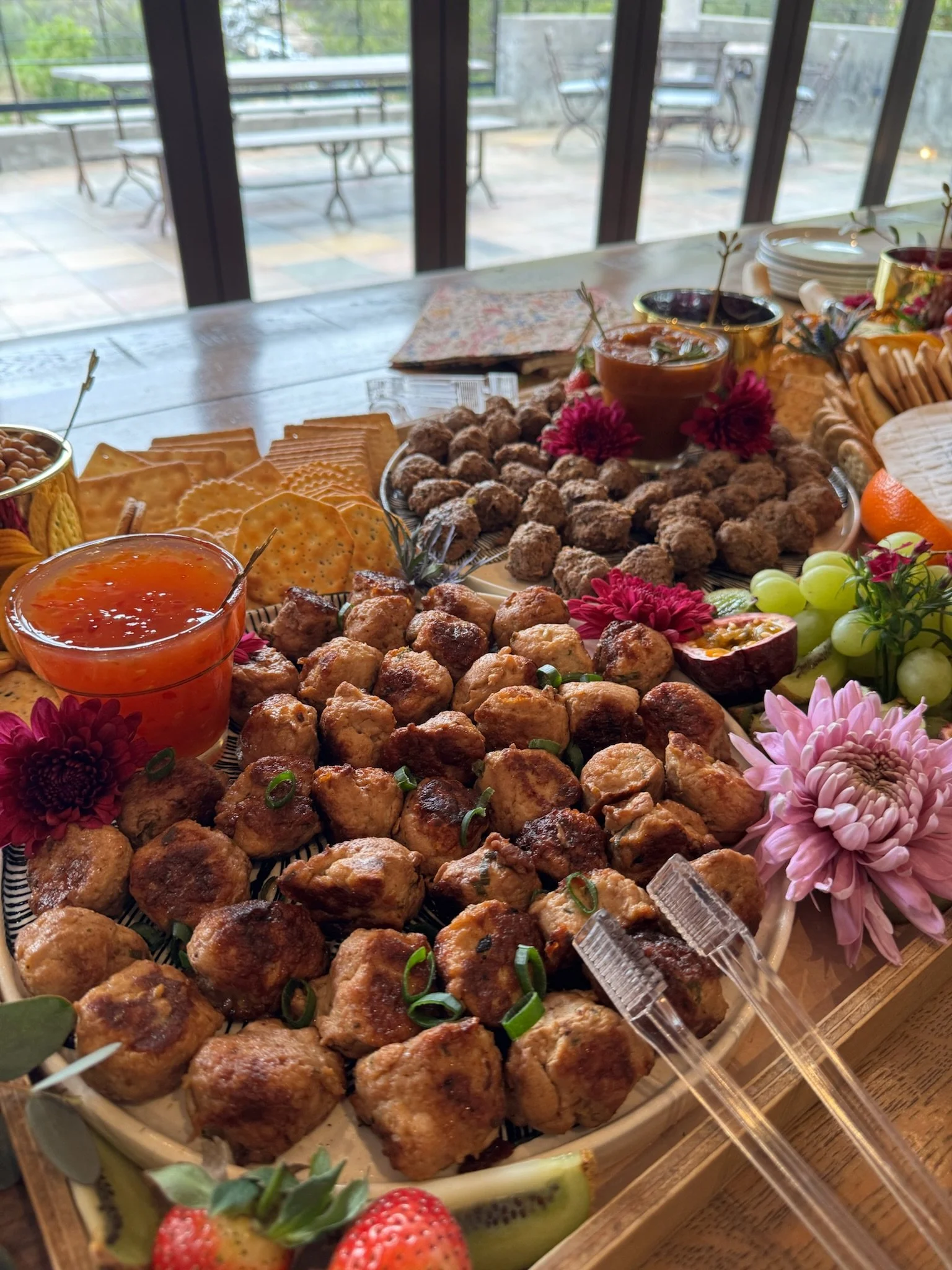 A table with a variety of appetizers, including meatballs, dipping sauces, cheese, grapes, strawberries, and flowers, set in front of large window doors.