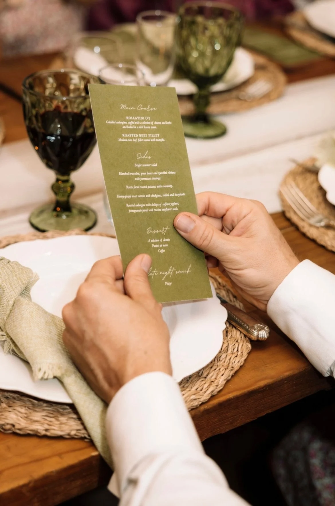 Person holding a green menu or program at a dining table set with plates, silverware, and glasses, including a glass of red wine.