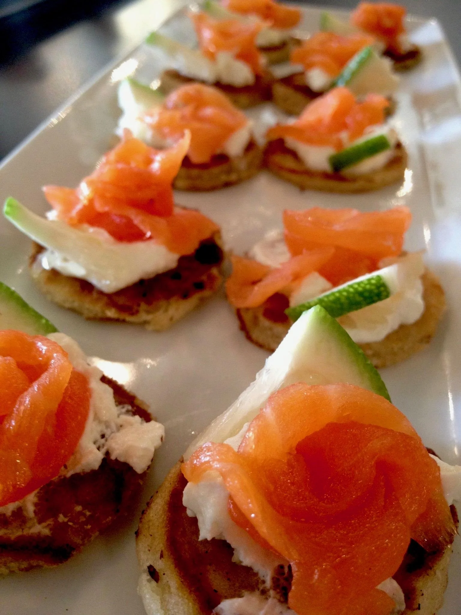 Close-up of smoked salmon canapés with cream cheese and cucumber slices on toasted bread rounds arranged on a white plate.