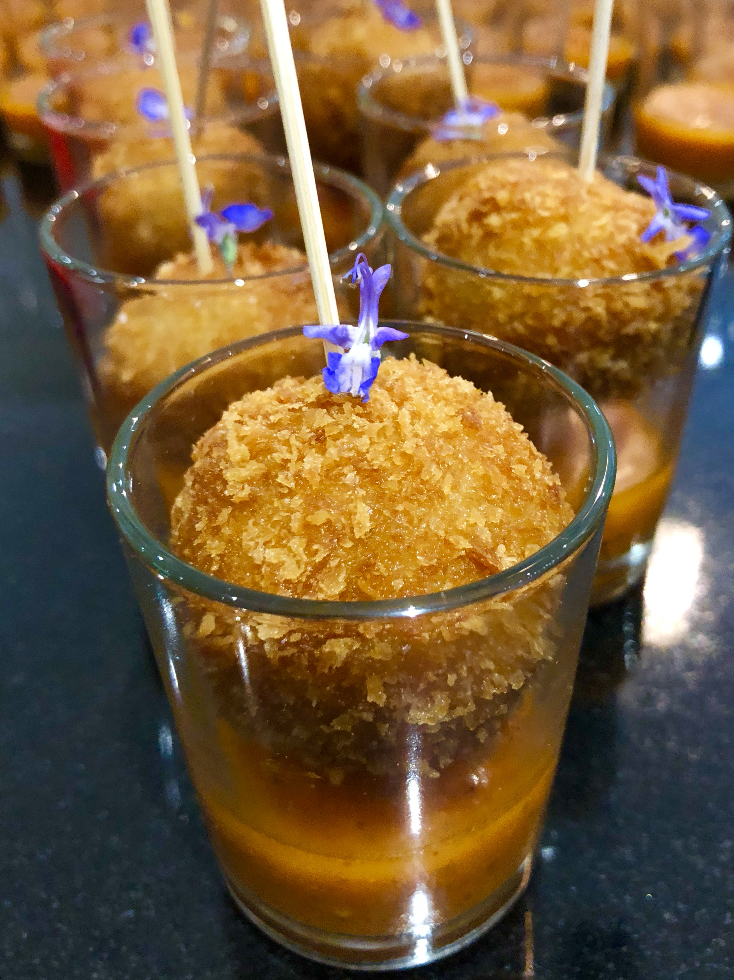 Multiple glasses with crispy fried balls topped with purple flowers and skewers on a black surface.
