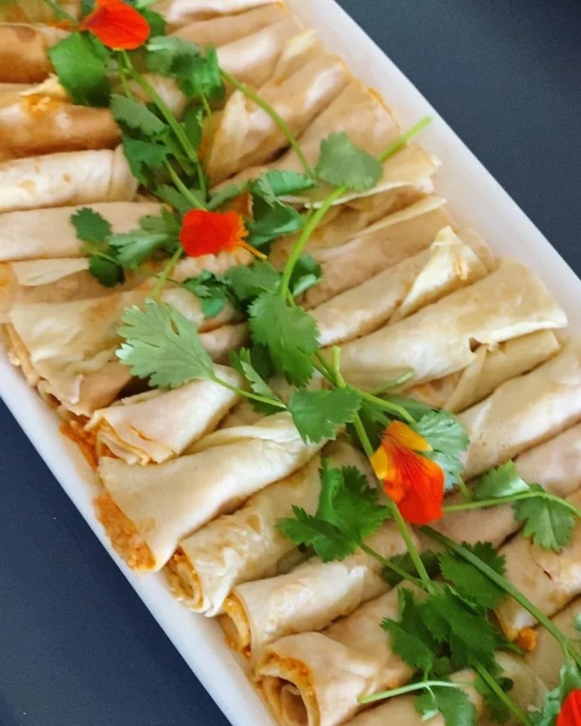 A white rectangular platter with neatly arranged rolled egg rolls garnished with fresh cilantro and orange edible flowers.