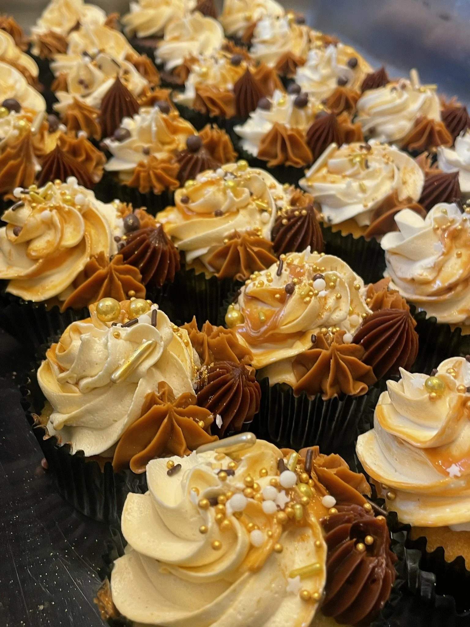 Assorted cupcakes decorated with swirls of frosting in caramel, vanilla, and chocolate flavors, topped with sprinkles, gold beads, and chocolate decorations.