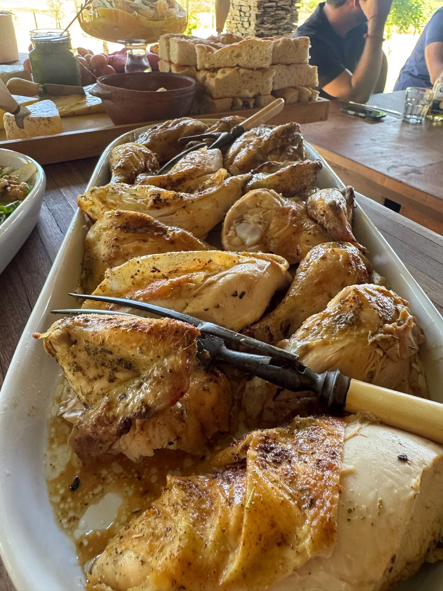 A large platter of cooked chicken pieces with golden-brown skin, garnished with herbs, and a carving fork resting on top. In the background, there are slices of bread stacked on a wooden cutting board, a jar of green sauce, and various condiments on 