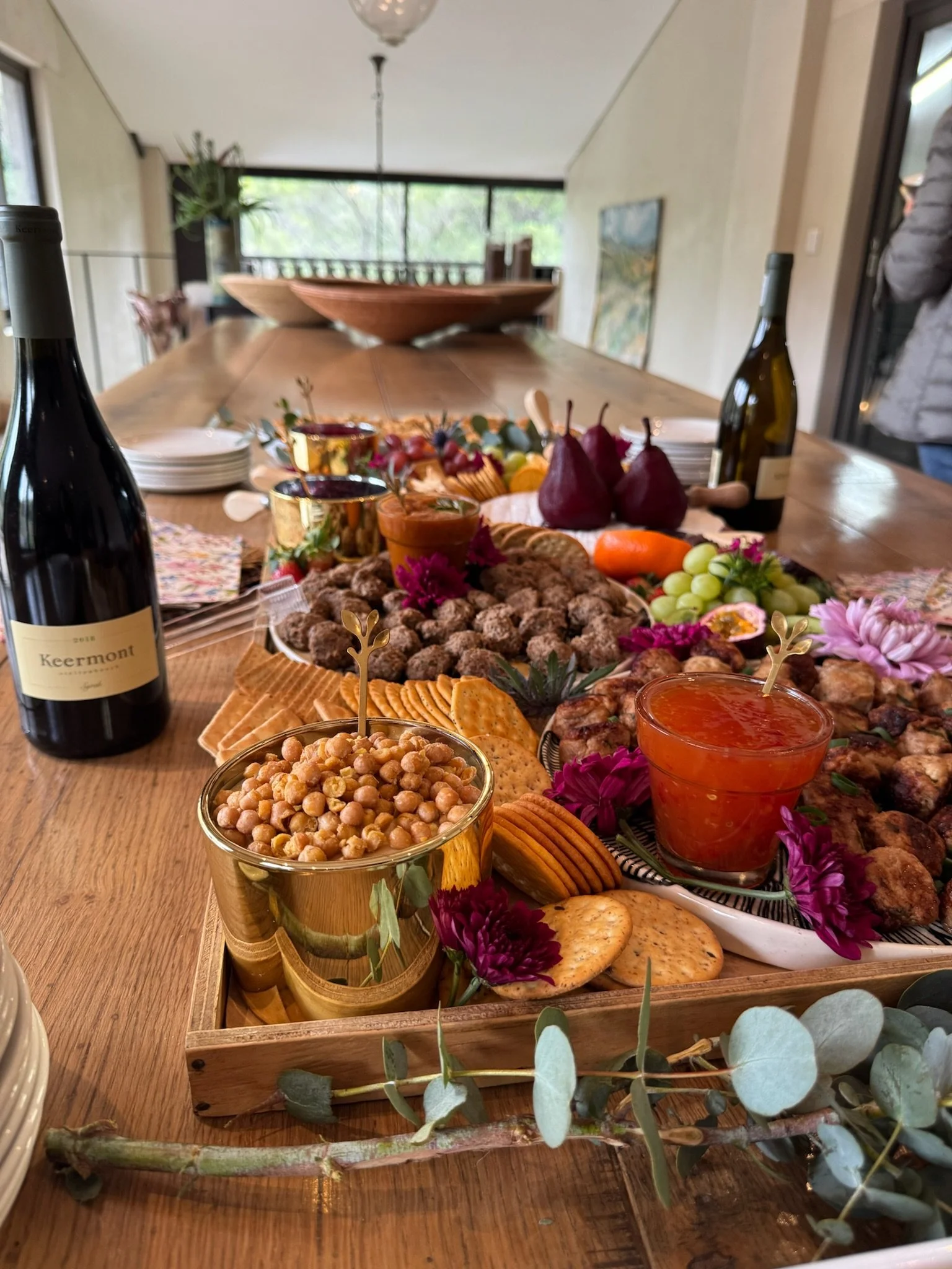 A table set for a celebration with wine bottles, a variety of snacks including chickpeas, crackers, grapes, vegetables, and small bites, garnished with flowers and greenery.