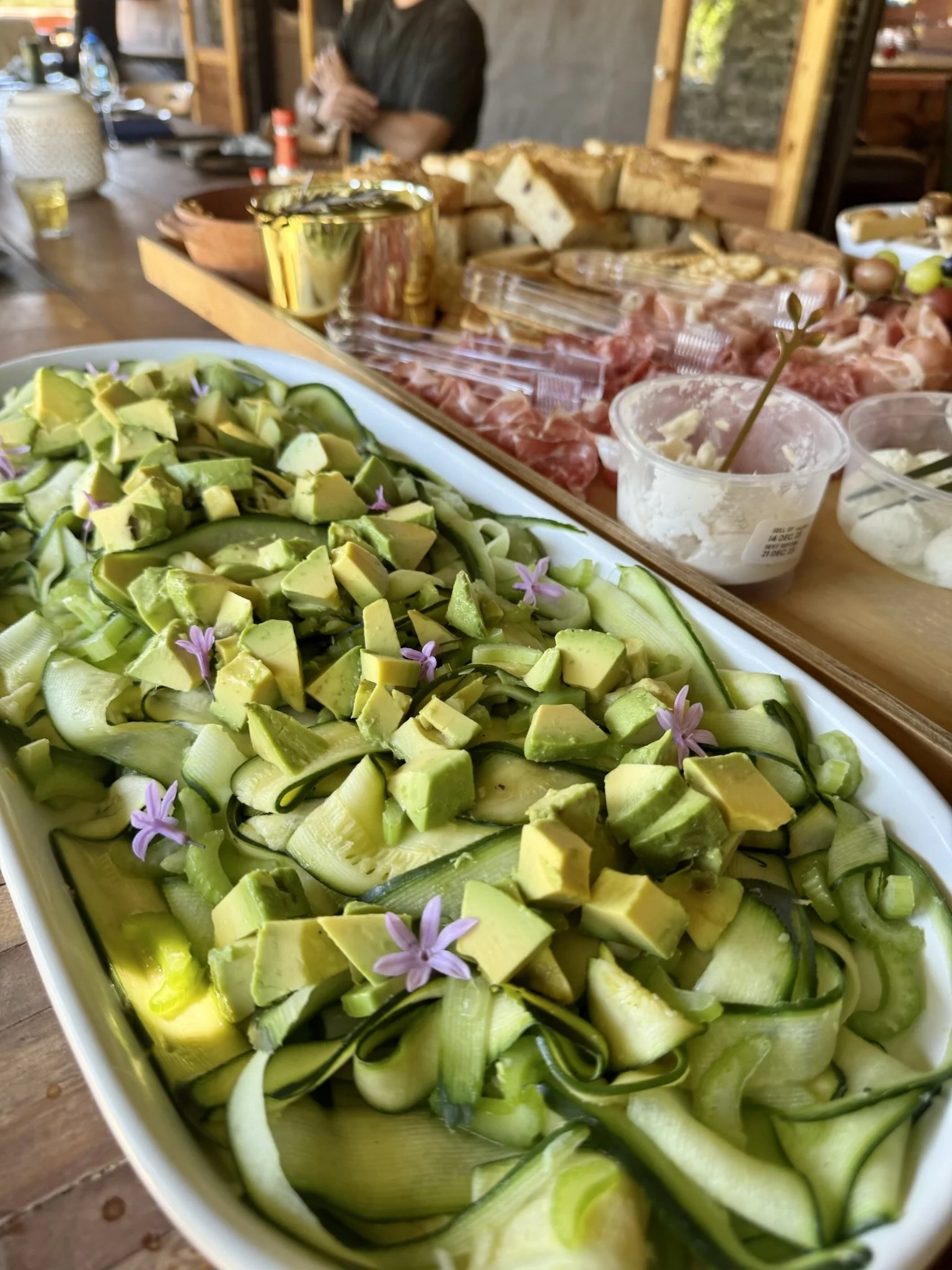 A plate of sliced zucchini topped with diced avocado and small purple flowers on a wooden table at a buffet.