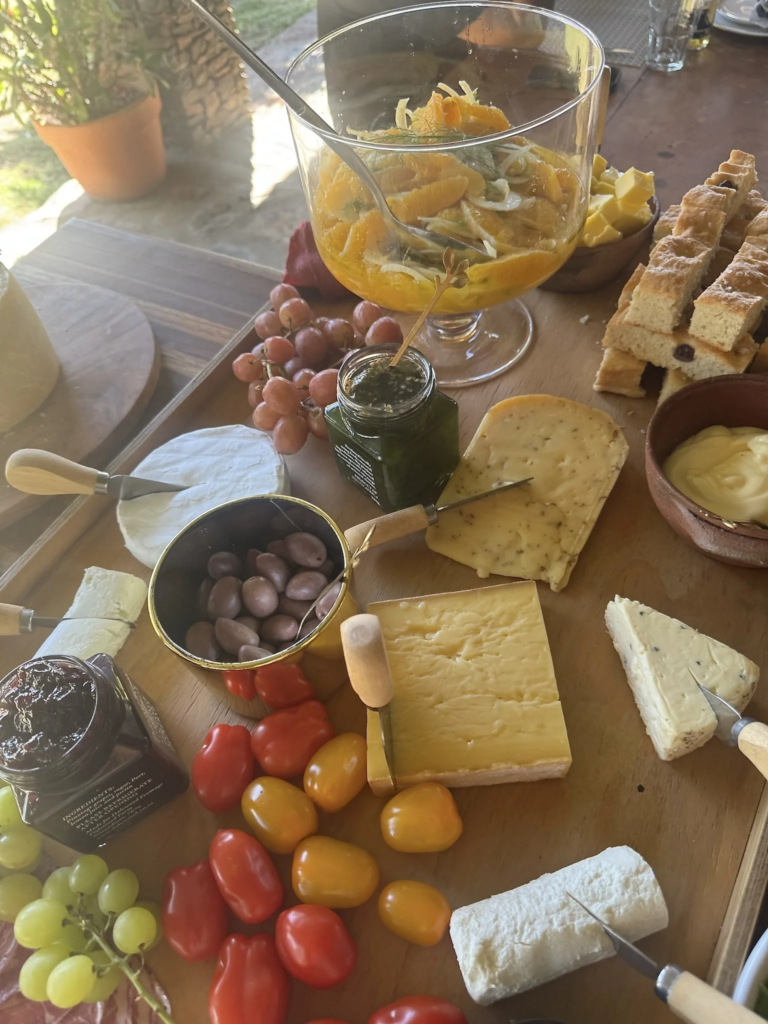 A cheese and fruit platter with various cheeses, grapes, cherry tomatoes, and a glass bowl of sliced oranges and lemons.