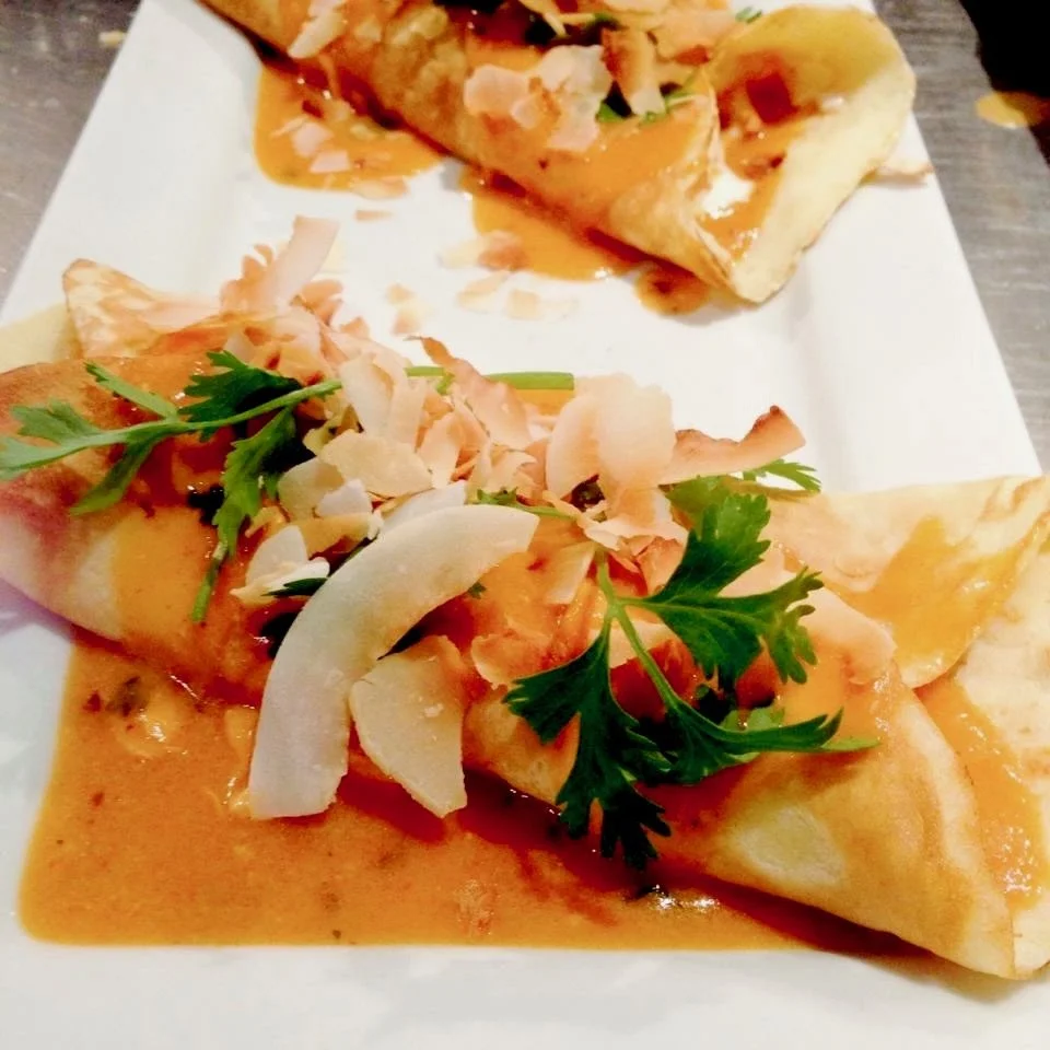 Close-up of two flavorful butter chicken crepes topped with cilantro, shredded coconut, and a creamy, spicy sauce, served on a white plate.