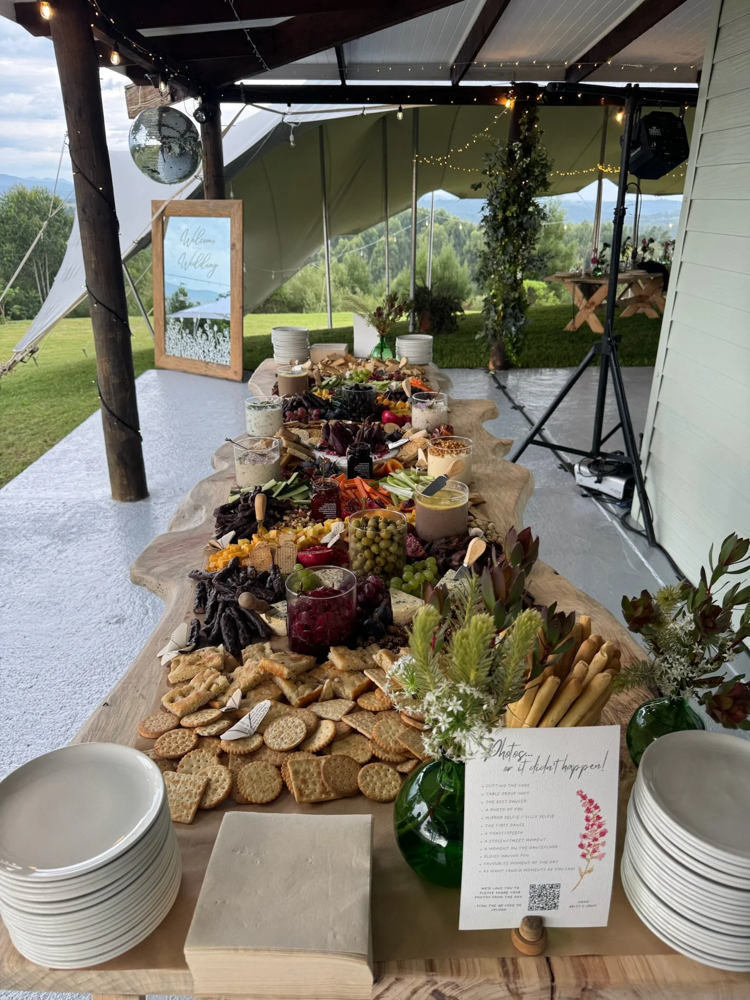 Table with an assortment of cheese, crackers, fruit, and snacks at an outdoor wedding reception under a pavilion. A chalkboard sign reads 'photos or it didn't happen!' with instructions. In the background, there are trees and mountains visible.