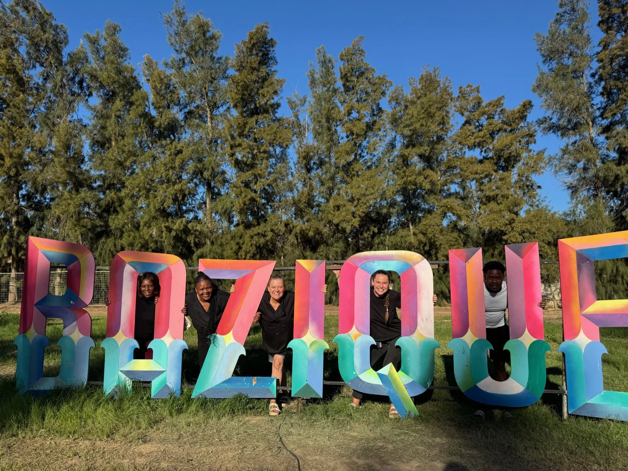 Five people are posing behind colorful 3D letters that spell 'BAZIQUE'. They are outdoors on grass with trees and a blue sky in the background.